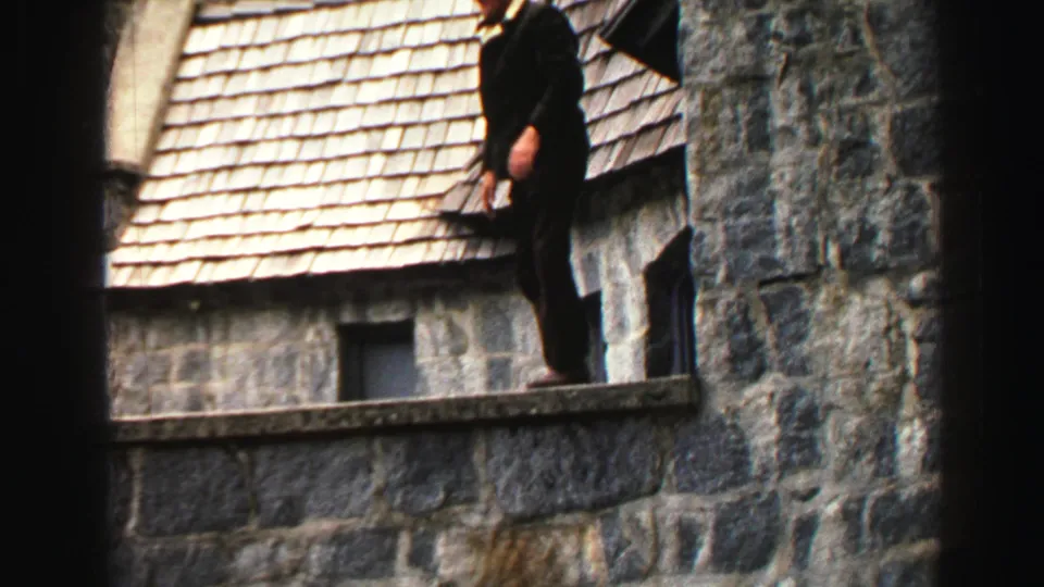 1969 Lake Tahoe home-movie frame showing a man in a dark suit standing on a stone ledge near a shingled roof and small window.
