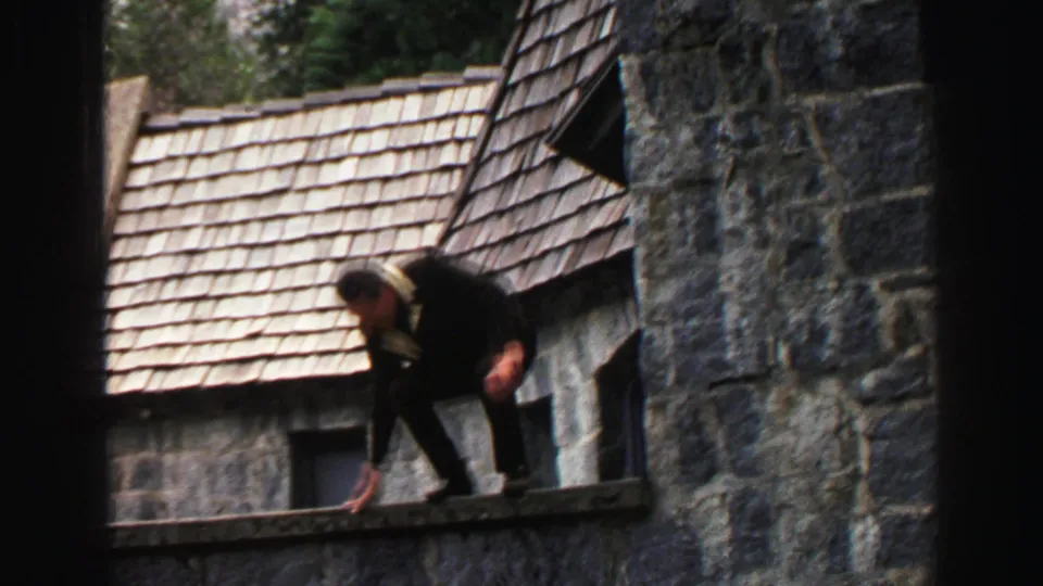 1969 Lake Tahoe home-movie frame showing the man crouching on a narrow ledge of a rustic stone building.