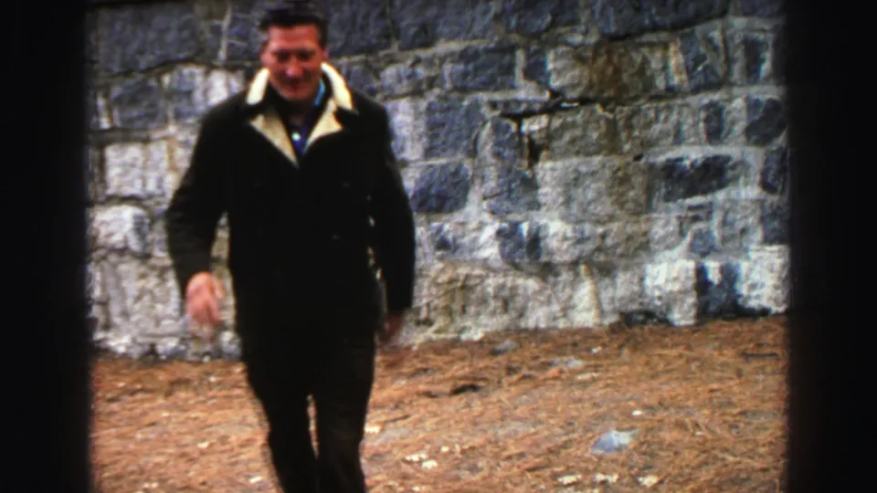 A smiling man approaches the camera along dirt ground beside a stone wall in Lake Tahoe home-movie footage.