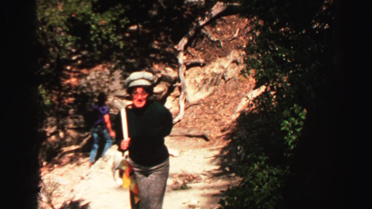 Hikers climb a rocky mountain trail, with a woman in a green sweater approaching the camera.