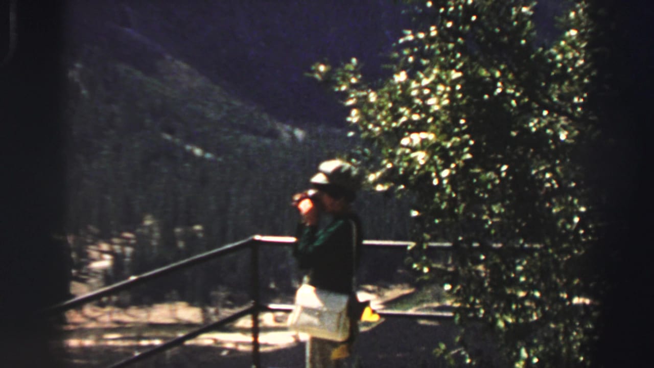 Woman in a hat stands by a railing and looks through binoculars.