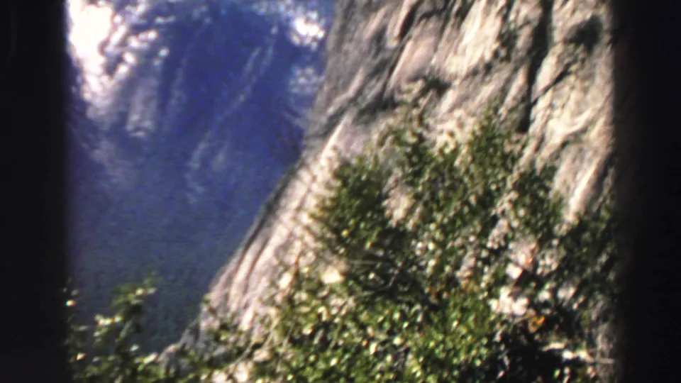 1969 Lake Tahoe-area frame showing a steep rocky slope, shrubs, and distant shaded mountain terrain.