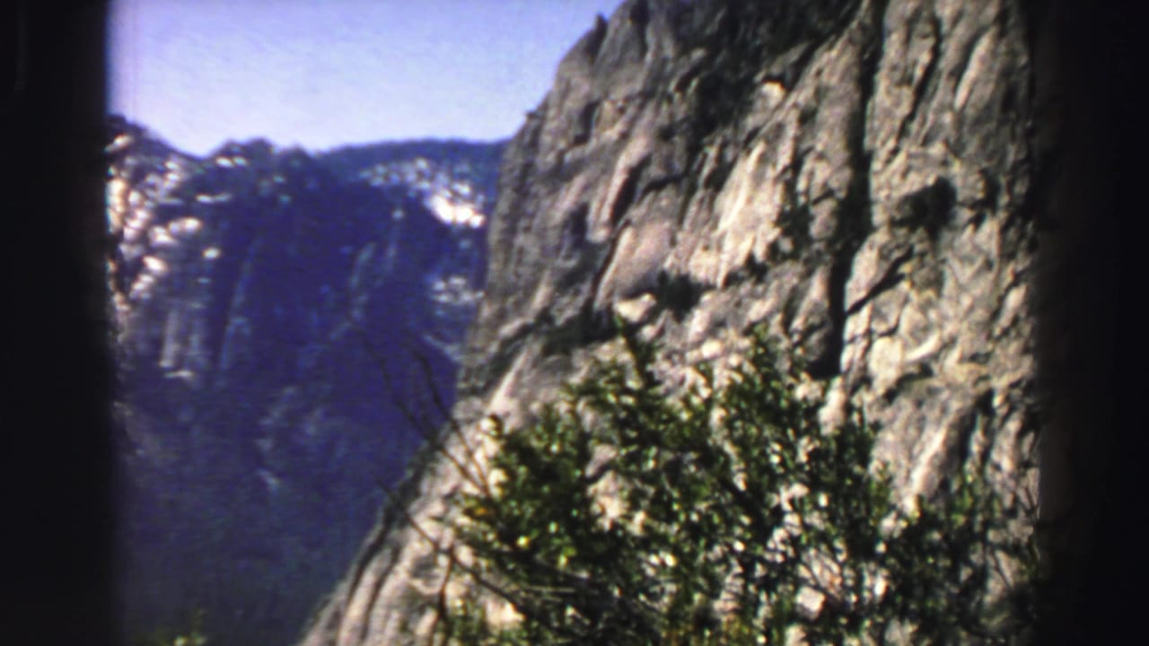 Rocky mountain cliff with distant snow patches, shrubs, and dark film-edge framing.