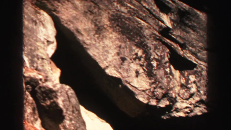 1969 home-movie frame of large granite boulders near Lake Tahoe, with warm sunlight, dark shadow pockets, and film grain.