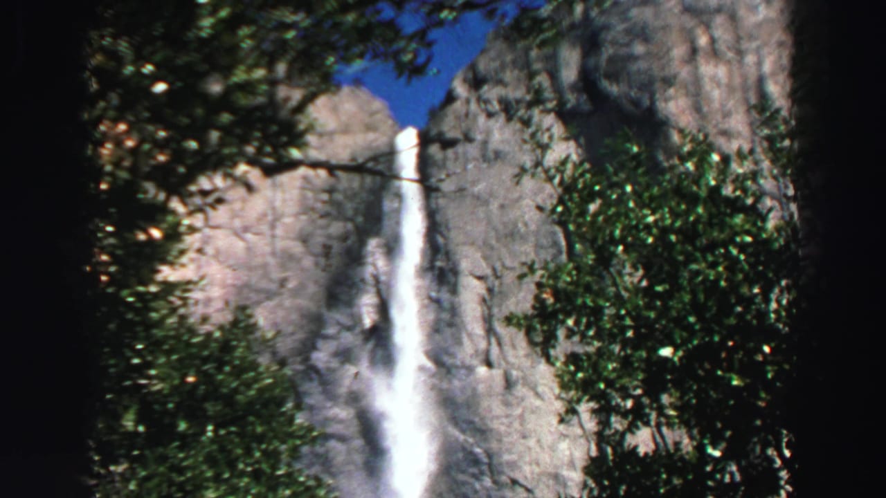 Waterfall drops down a granite cliff behind trees in bright sunlight.