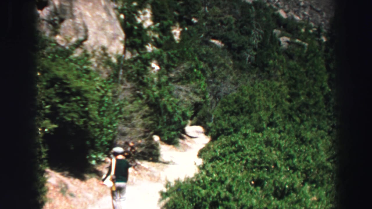 Distant hiker walking on a dirt trail through green forest and rocky terrain.
