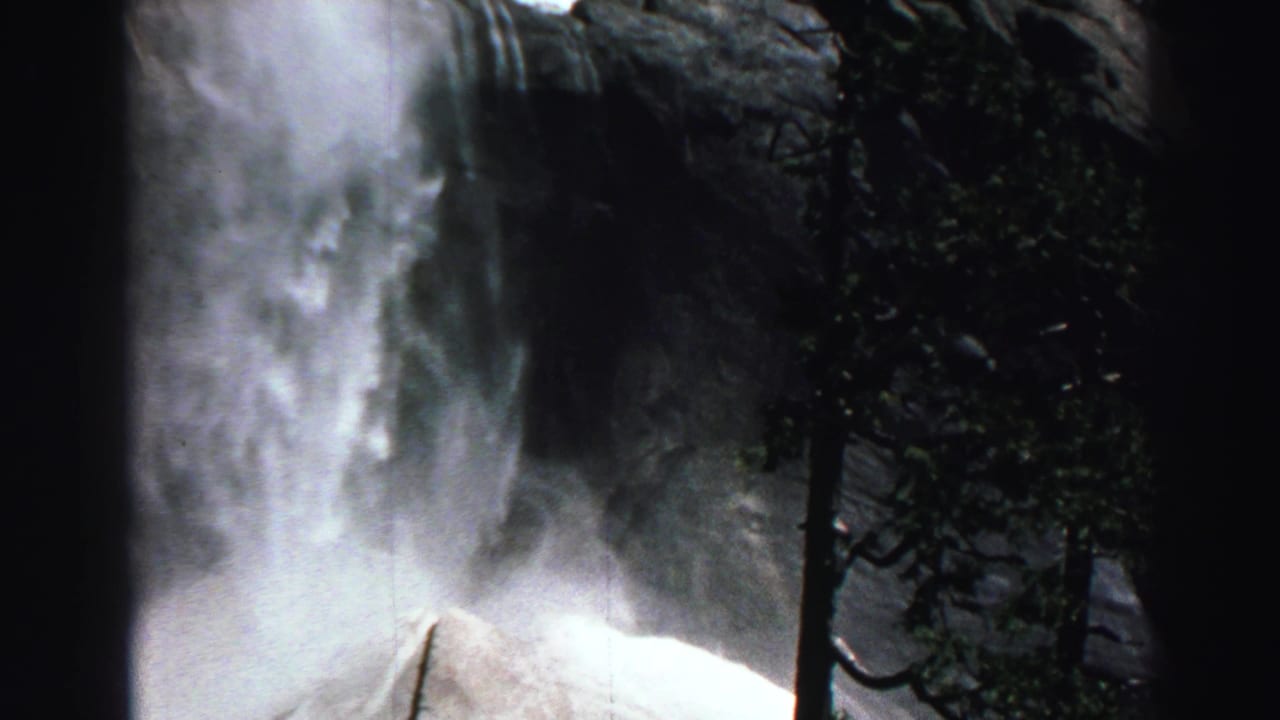 Waterfall mist and evergreen trees against a rocky cliff.