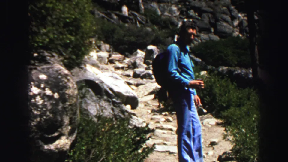 Adult hiker in a blue shirt and jeans stands on a rocky Lake Tahoe-area mountain trail with shrubs and granite around him.