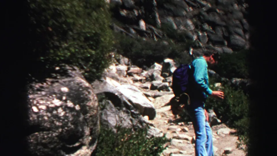 Backpacked adult hiker in blue clothing walks along an uneven rocky trail near Lake Tahoe, surrounded by shrubs and granite stones.