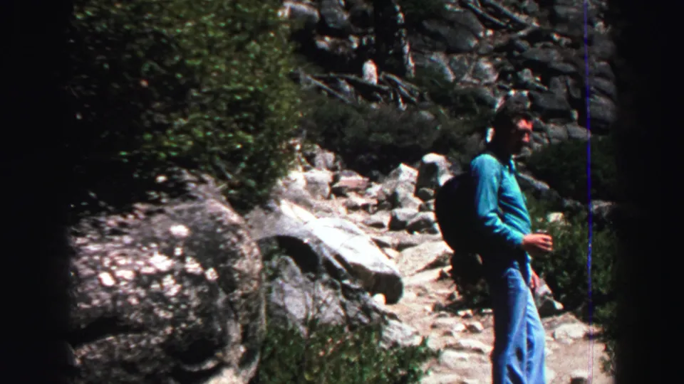 Adult hiker in a blue shirt and jeans stands on the Lake Tahoe-area trail, with backpack, shrubs, rocks, and dark 8mm frame edges visible.