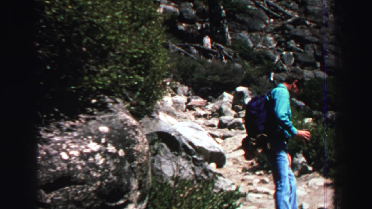 Adult man with backpack walking on a rocky mountain trail beside shrubs and boulders.