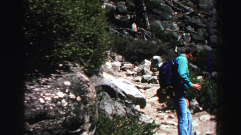 In 1969 Lake Tahoe footage, an adult man in a turquoise shirt and jeans walks along a rocky trail with shrubs and boulders nearby.