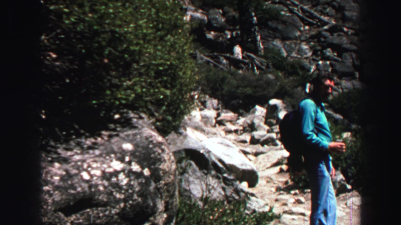In the Lake Tahoe home-movie scene, an adult man pauses or turns on a rocky mountain trail bordered by shrubs and large stones.
