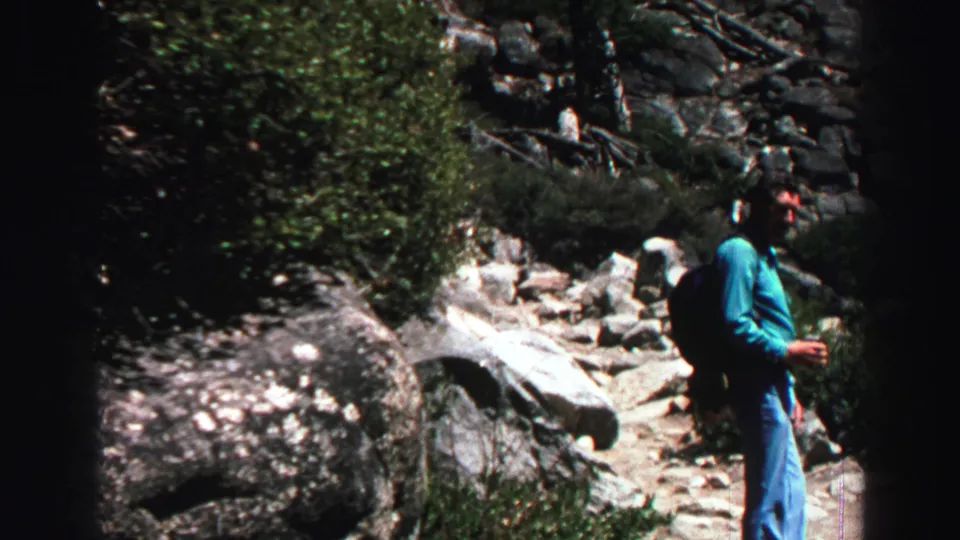 In the Lake Tahoe home-movie scene, an adult man pauses or turns on a rocky mountain trail bordered by shrubs and large stones.