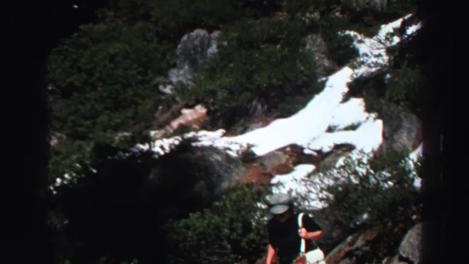 Lake Tahoe mountain terrain shows lingering snow across rocks and shrubs, with the top of an adult hiker partly visible near the lower frame edge.