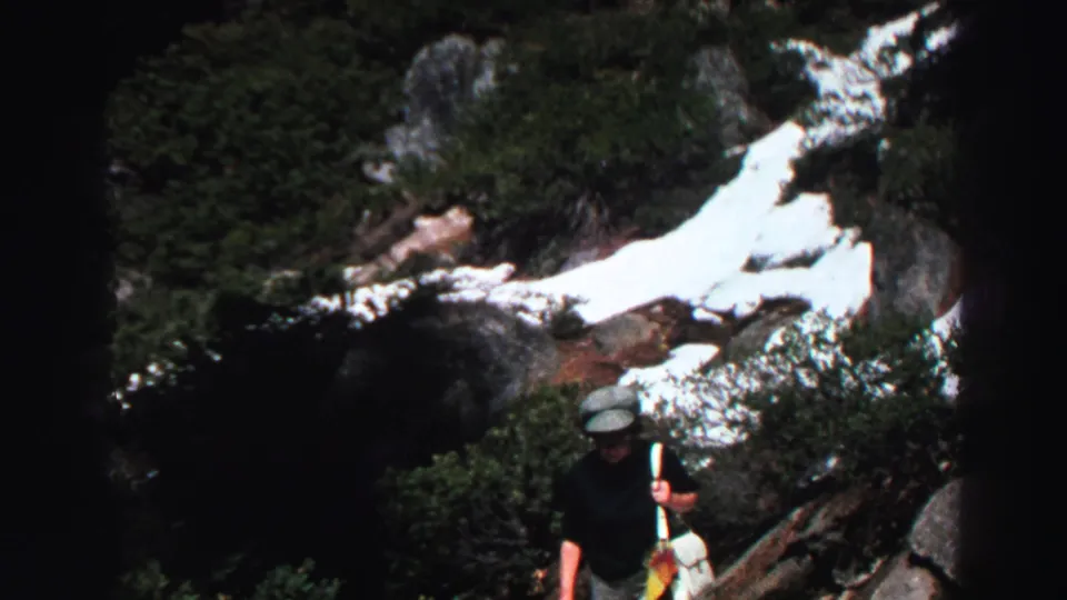 An adult woman in a hat moves through the Lake Tahoe mountain trail scene, carrying a bag and walking stick below snow-streaked rocks.