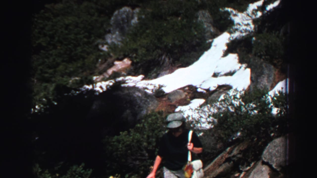 Hiker in cap and dark shirt climbs through rocks, shrubs, and snow patches.
