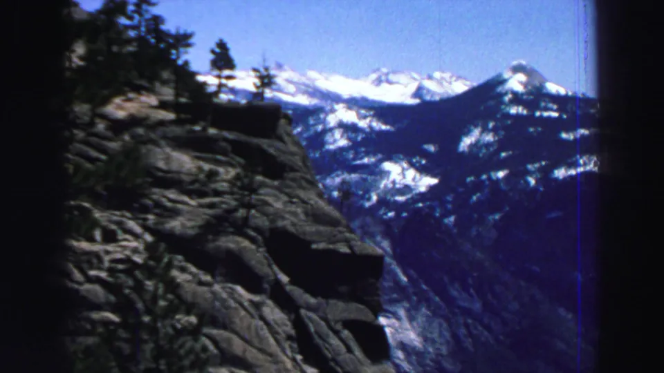A rocky Lake Tahoe-area mountain overlook frames evergreen trees and snow-capped Sierra Nevada peaks under blue sky.