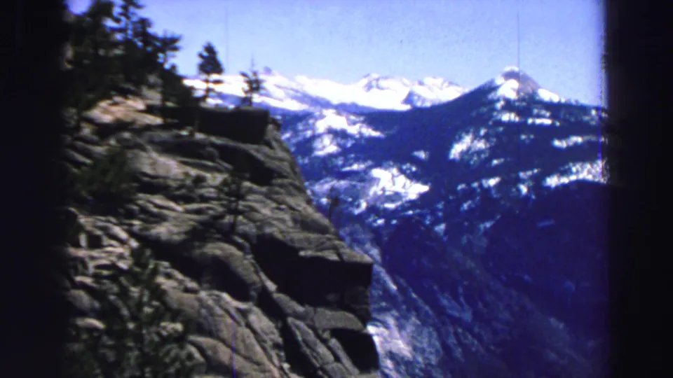 Handheld 8mm view of a rugged rocky slope, sparse evergreens, and distant snow-covered mountain ridges near Lake Tahoe.