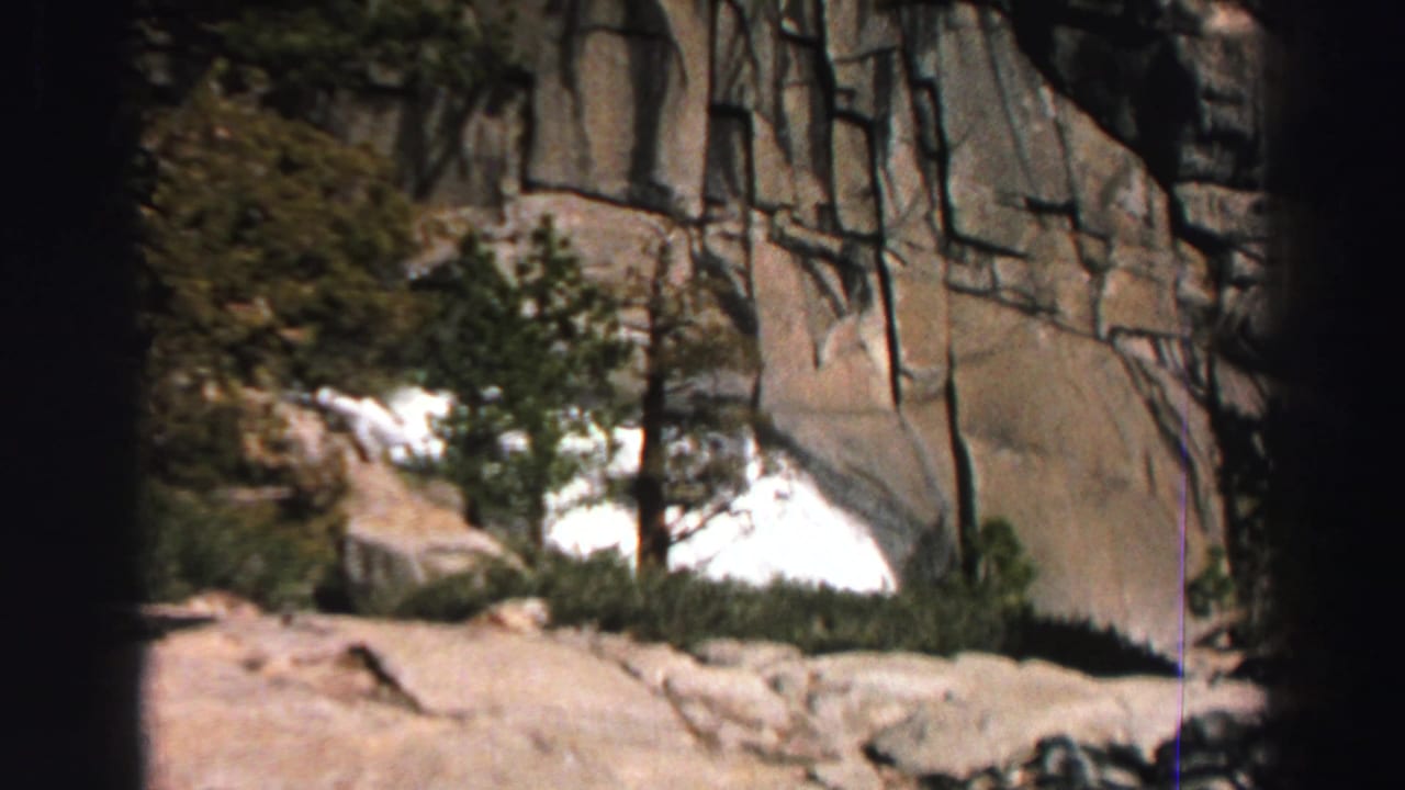 1969 Lake Tahoe 8mm frame showing granite rock, evergreens, and a bright patch of water or snow in a mountain setting.