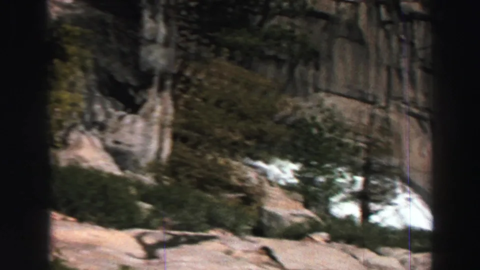 1969 Lake Tahoe 8mm frame looking across evergreens, granite cliffs, and a bright white water or snow patch in the landscape.