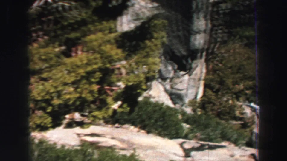 1969 Lake Tahoe home-movie frame showing pale mountain rock, shrubs, and evergreen trees from an elevated viewpoint.