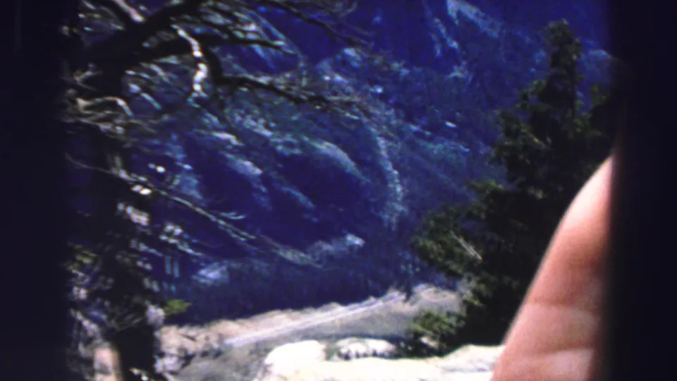 Handheld 1969 Lake Tahoe frame with blue mountain slopes, evergreens, pale rocks, and a hand entering the view.
