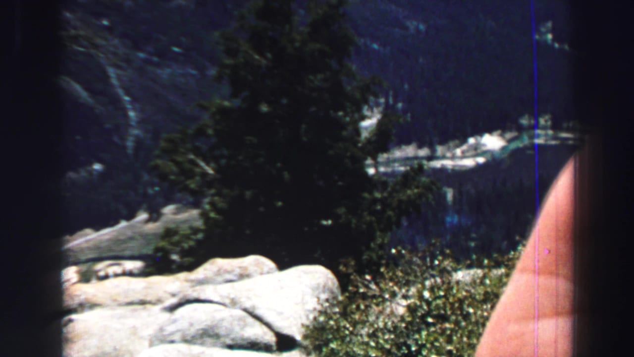 Forested mountain slope, granite rocks, lake water, and a partial hand at the frame edge.