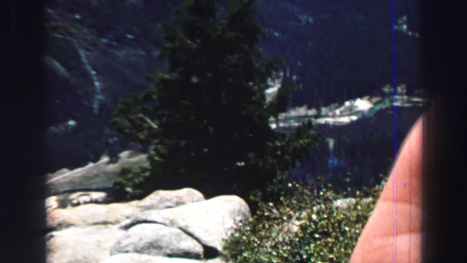 1969 Lake Tahoe home-movie frame showing trees, pale rocks, dark lake water, and a partial hand or arm at the right edge.