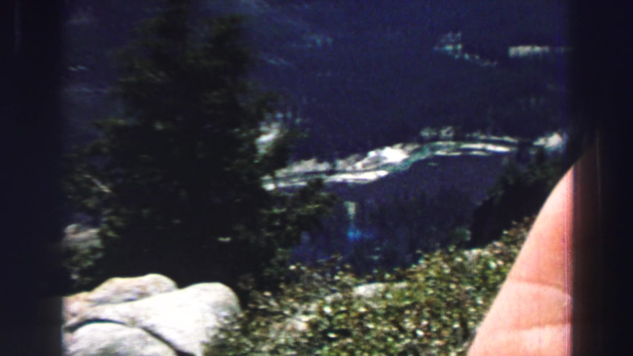 Mountain lake view with trees, rocks, shoreline, and a partial hand or arm on the right.