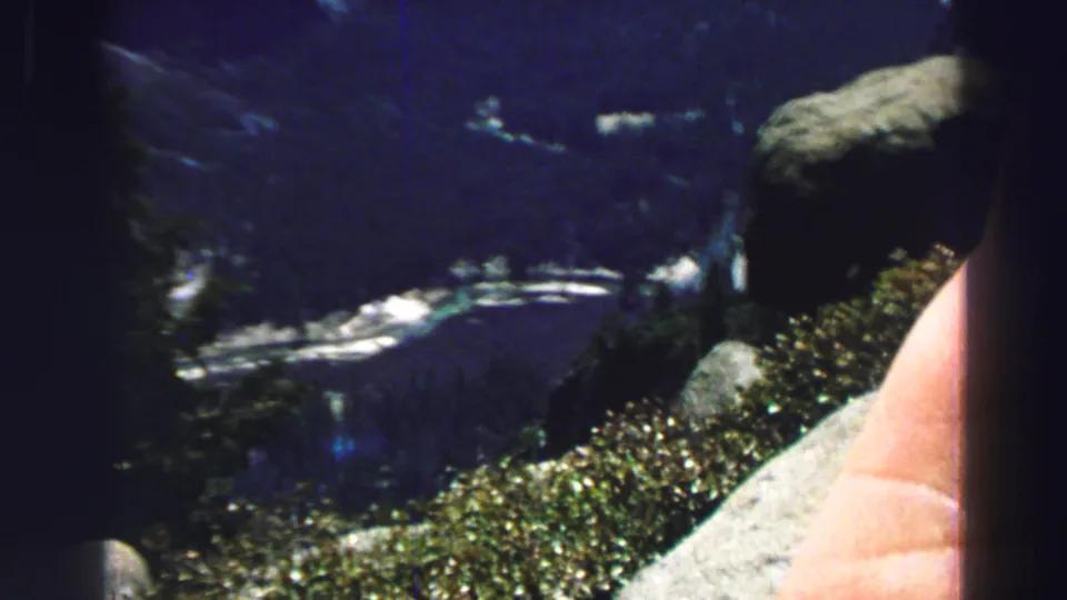 1969 Lake Tahoe scenic frame with dark water, distant wooded terrain, foreground shrubs, rocks, and a partial human arm near the lens.