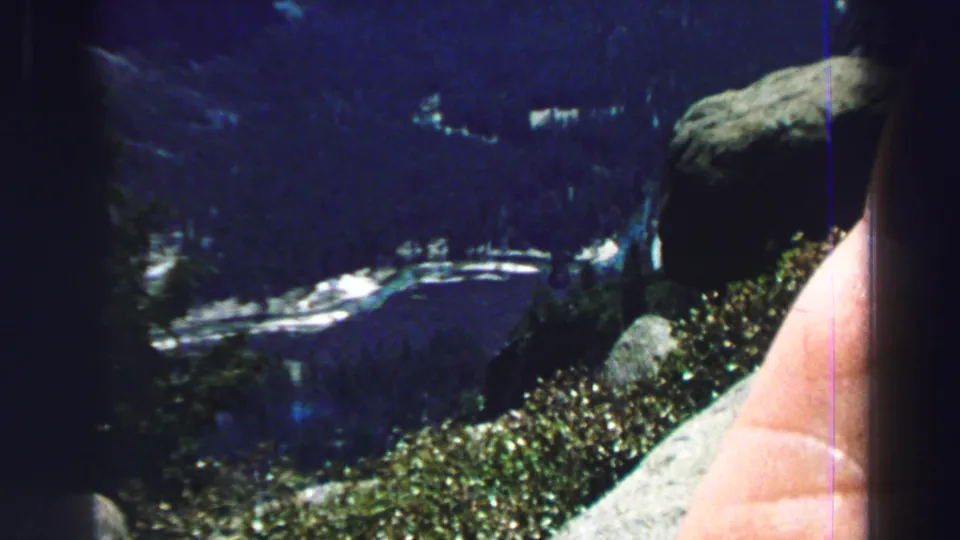 1969 8mm Lake Tahoe frame showing a rocky overlook, vegetation, distant shoreline, dark lake water, and a partial hand or arm.