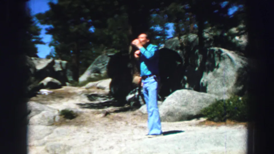1969 Lake Tahoe home-movie frame showing a man in blue flared jeans drinking from a cup among boulders and pine trees.