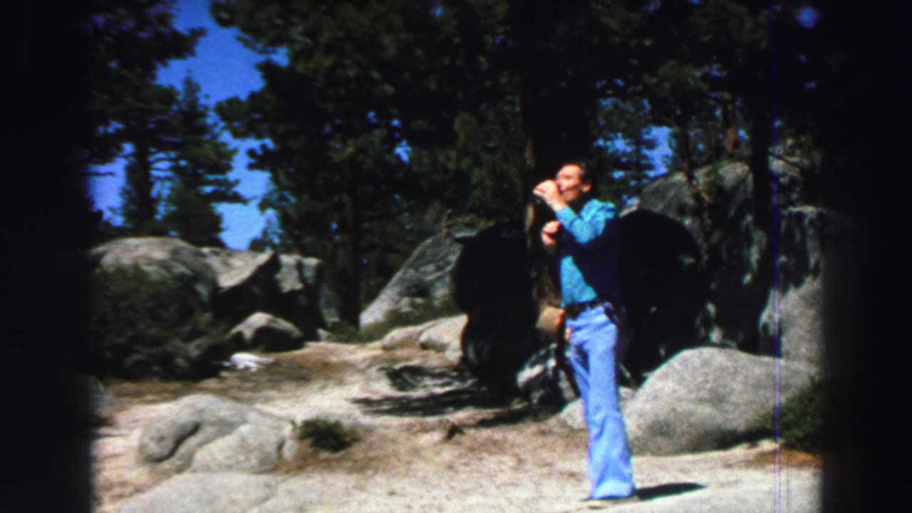 Man stands with a cup near large rocks and trees in bright sun.