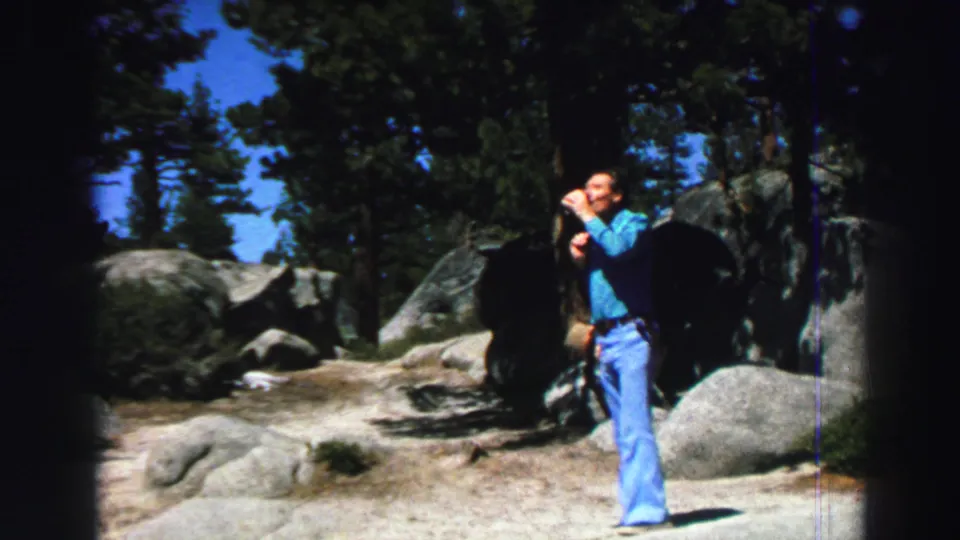1969 Lake Tahoe frame of a man holding a cup on a sunlit rocky path with pine trees and granite boulders behind him.