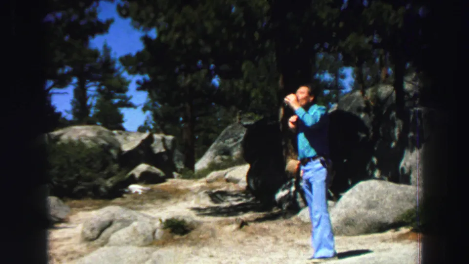 1969 Lake Tahoe home-movie still showing the man drinking on a sandy trail bordered by large rocks and pine trees.