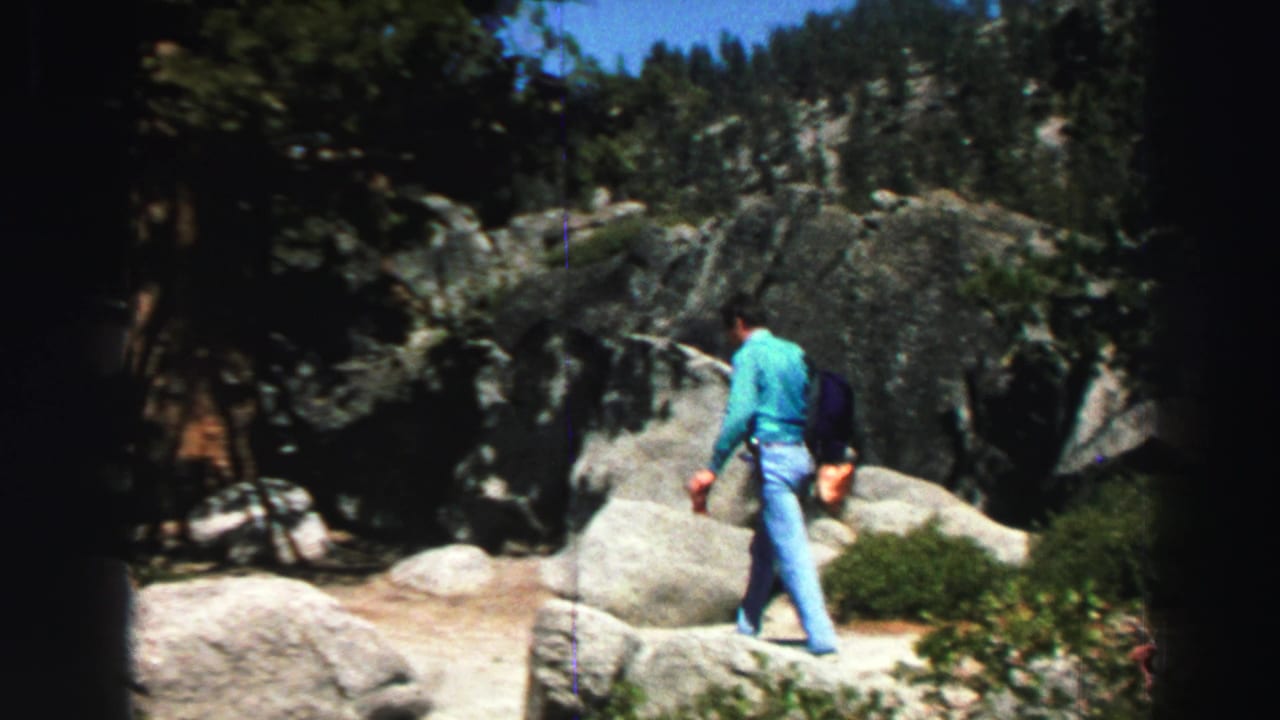In supplied 1969 Lake Tahoe home-movie footage, a man in a turquoise shirt and jeans hikes across sunlit granite boulders beneath pine trees.