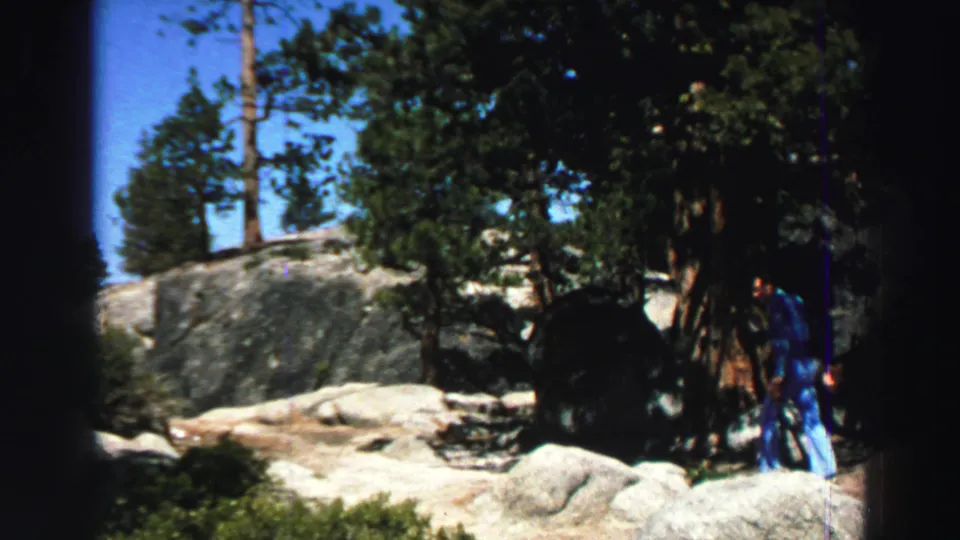 A rocky pine clearing fills the frame as the man appears partly at the right edge near a large tree in the Lake Tahoe home-movie scene.