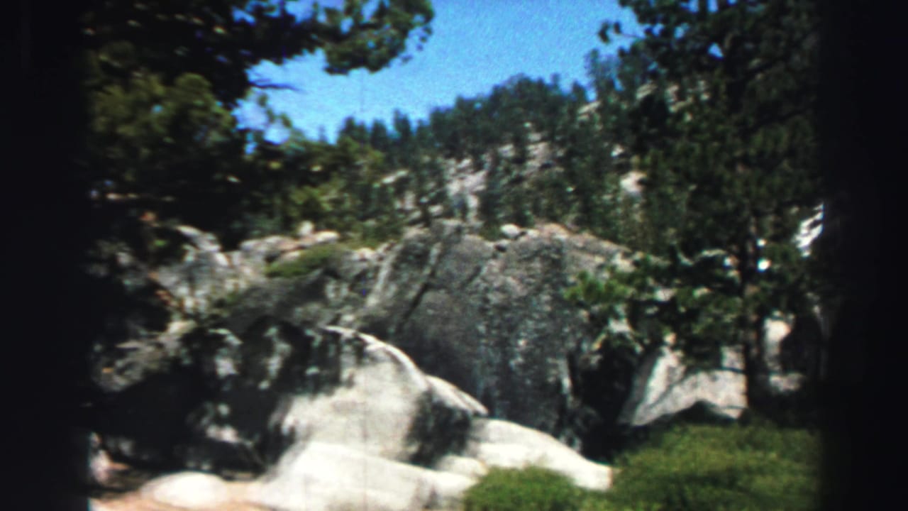 1969 Lake Tahoe home-movie frame showing pale granite boulders, evergreen trees, green ground cover, and blue sky.