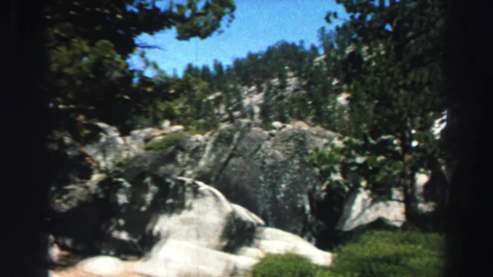 1969 Lake Tahoe home-movie frame showing pale granite boulders, evergreen trees, green ground cover, and blue sky.