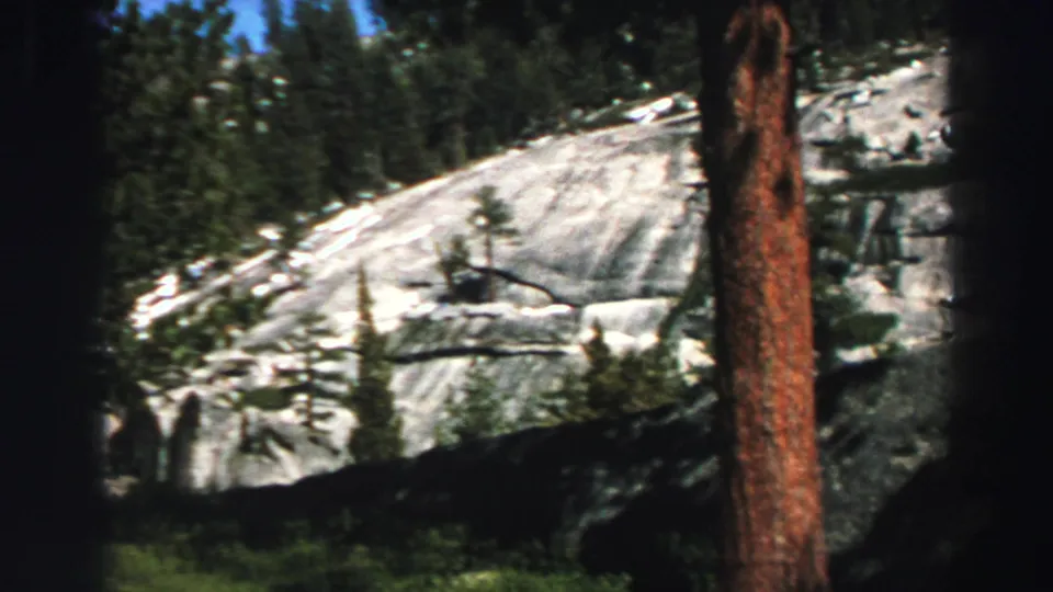 1969 Lake Tahoe home-movie view with reddish tree trunks in front of a pale granite hillside and evergreen forest.