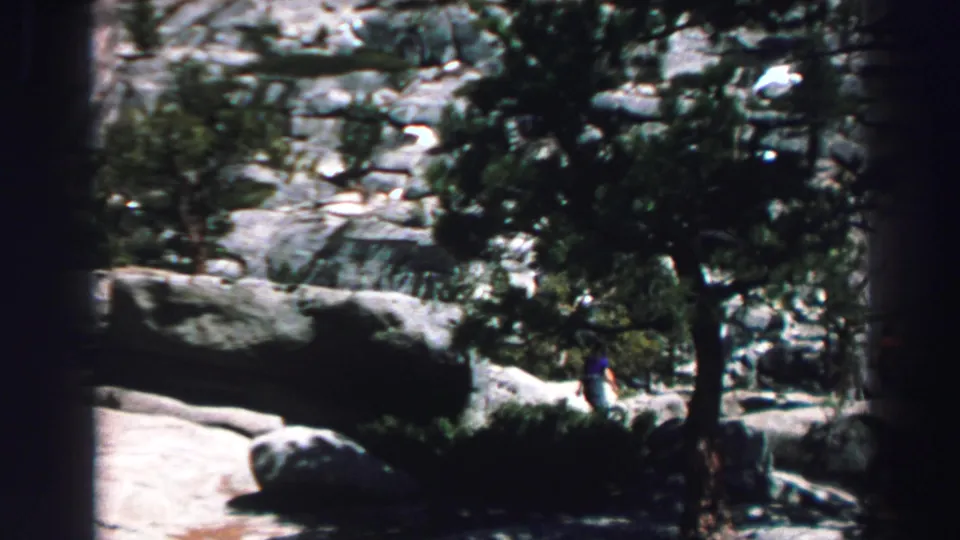1969 Lake Tahoe home-movie frame showing a distant person in blue near pine trunks and pale granite boulders.