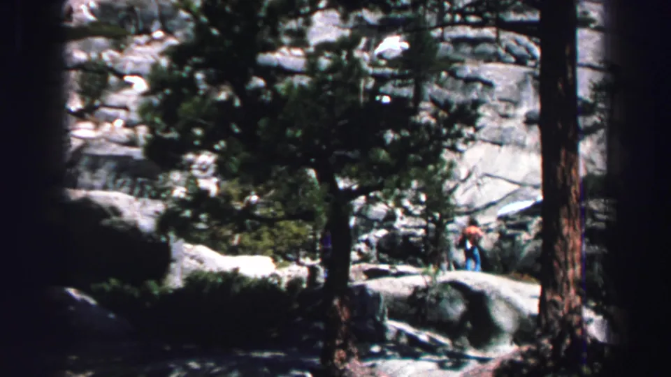 Lake Tahoe 1969 home-movie frame showing two distant people navigating boulders and pine shade on a forest trail.