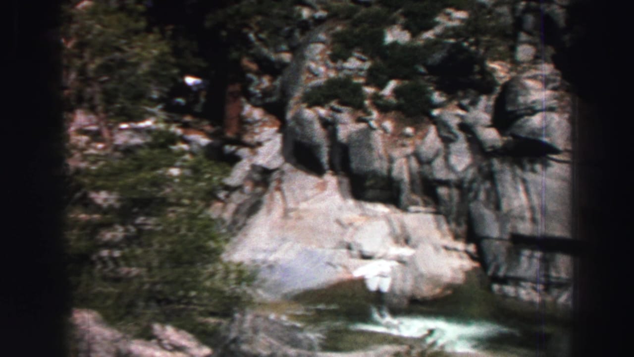 Steep rock formations, evergreen trees, and a green stream pool seen through a narrow film frame.