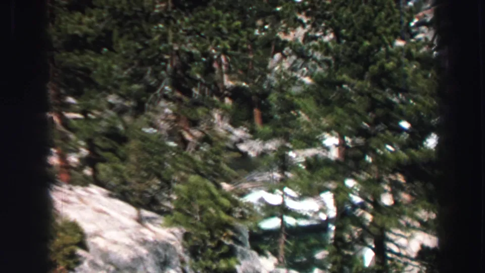 Handheld 8mm frame of Lake Tahoe terrain with dense evergreens, pale rock, snow pockets, and water visible below.