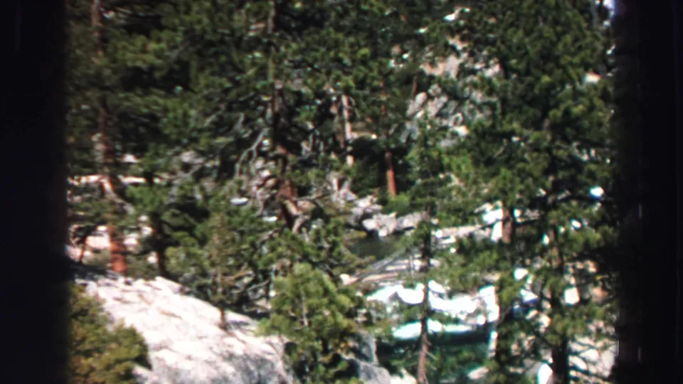 1969 Lake Tahoe frame with dense evergreens, exposed rock, patches of snow, and turquoise water glimpsed through the hillside.