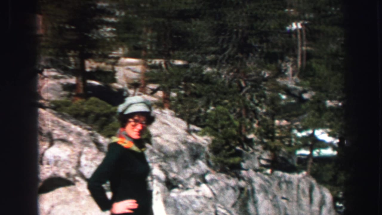 Woman in a hat posing on rocky ground among pine trees.