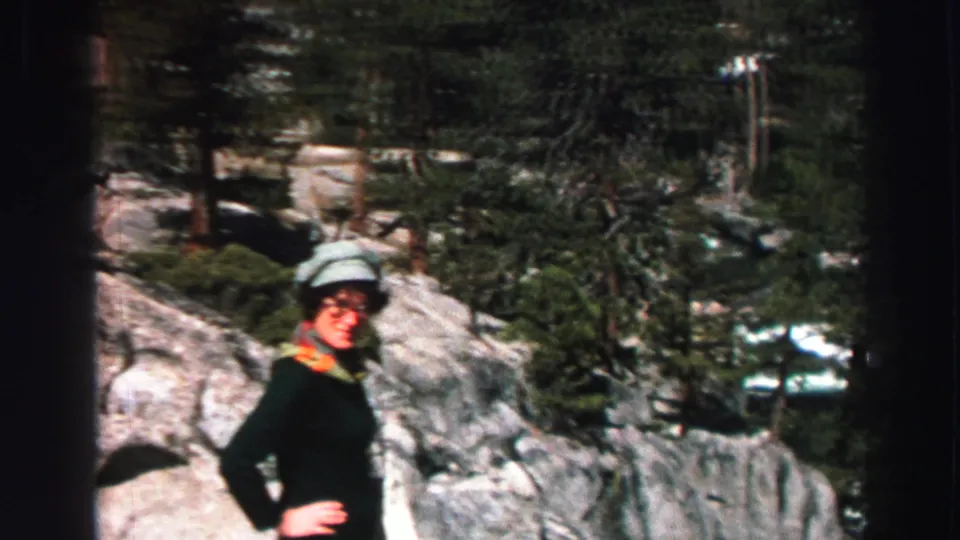 1969 Lake Tahoe home-movie frame showing a woman in a light hat standing on granite rocks near pine trees.