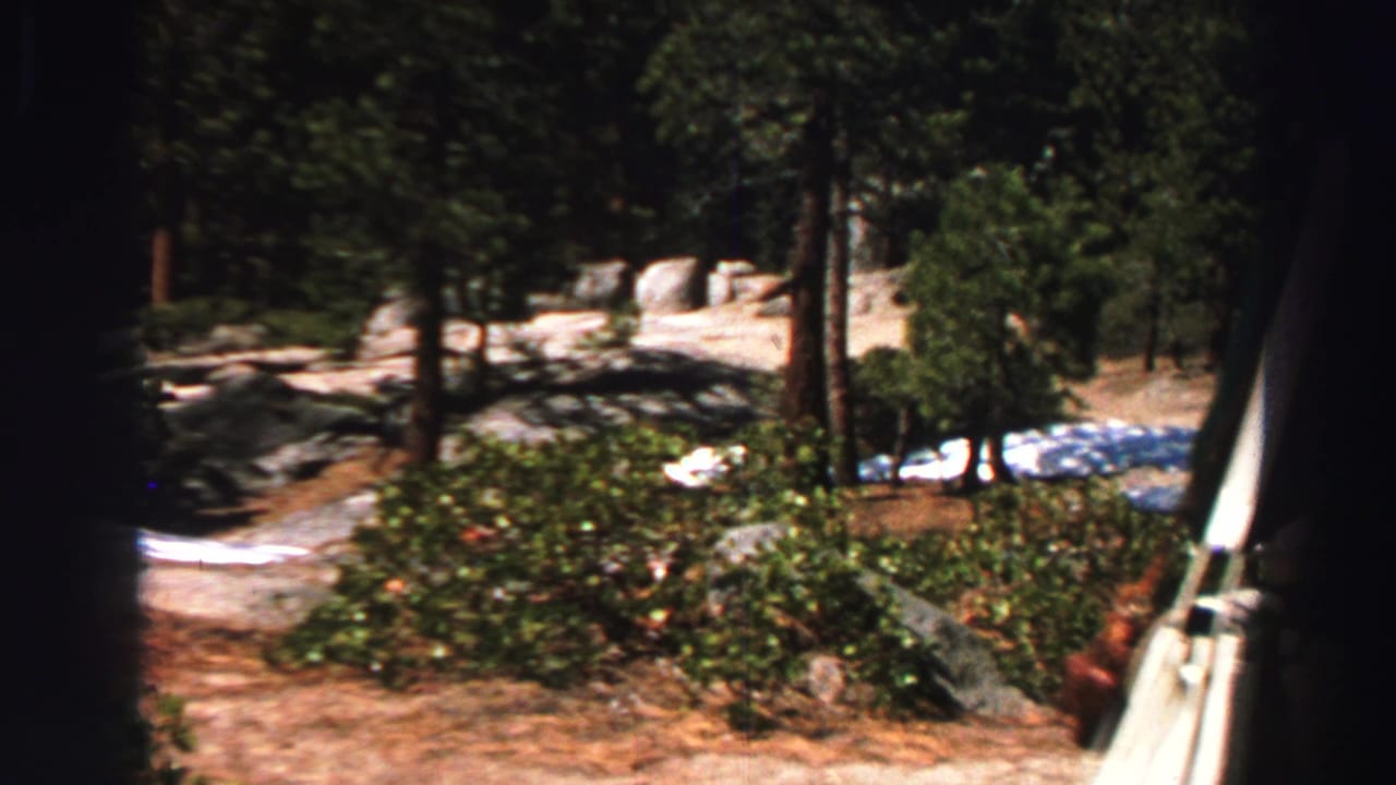 Lake Tahoe forest campsite view with granite rocks, pines, shrubs, small snow patches, and vehicles in the distance.