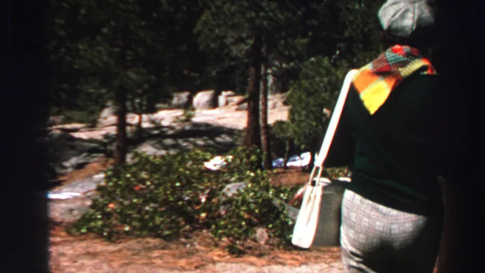 A woman with a shoulder bag walks through a Lake Tahoe pine and granite campsite in 1969 home-movie footage.